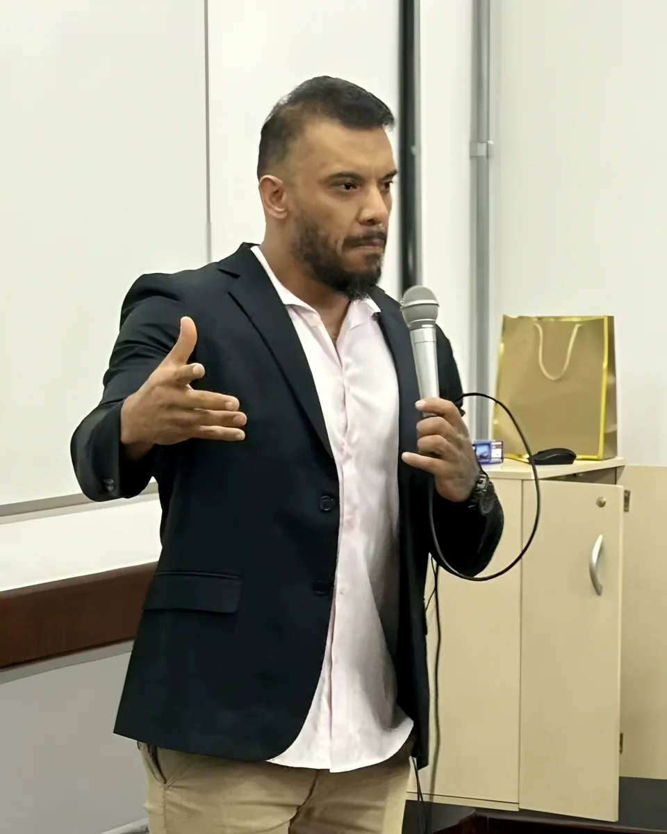 Alexsandro Rubin de blazer, com microfone, palestrando em uma sala de aula da faculdade